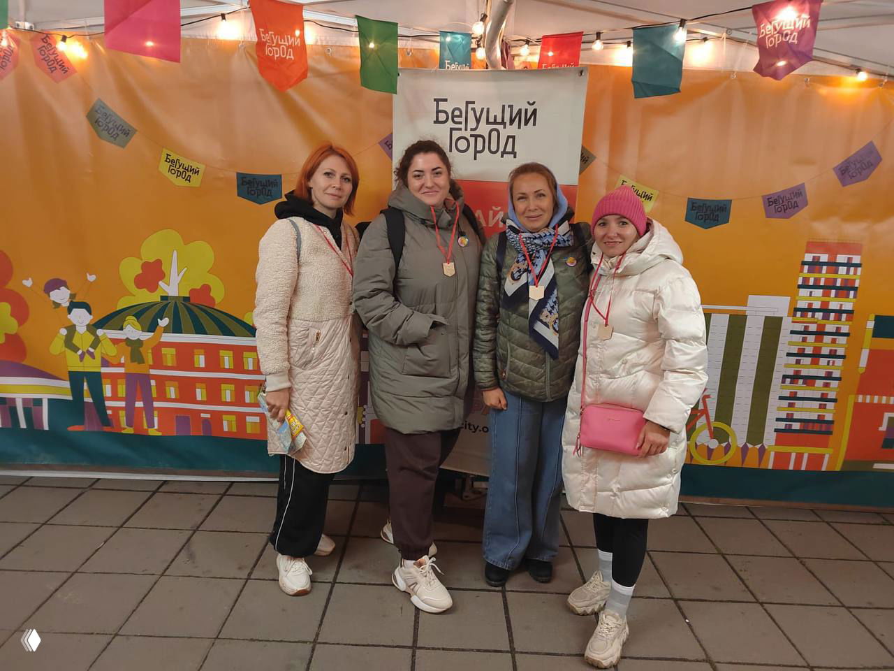 Четыре женщины стоят у стенда с логотипом «Бегущий город», на груди медали — команда после городского события или игры на свежем воздухе.