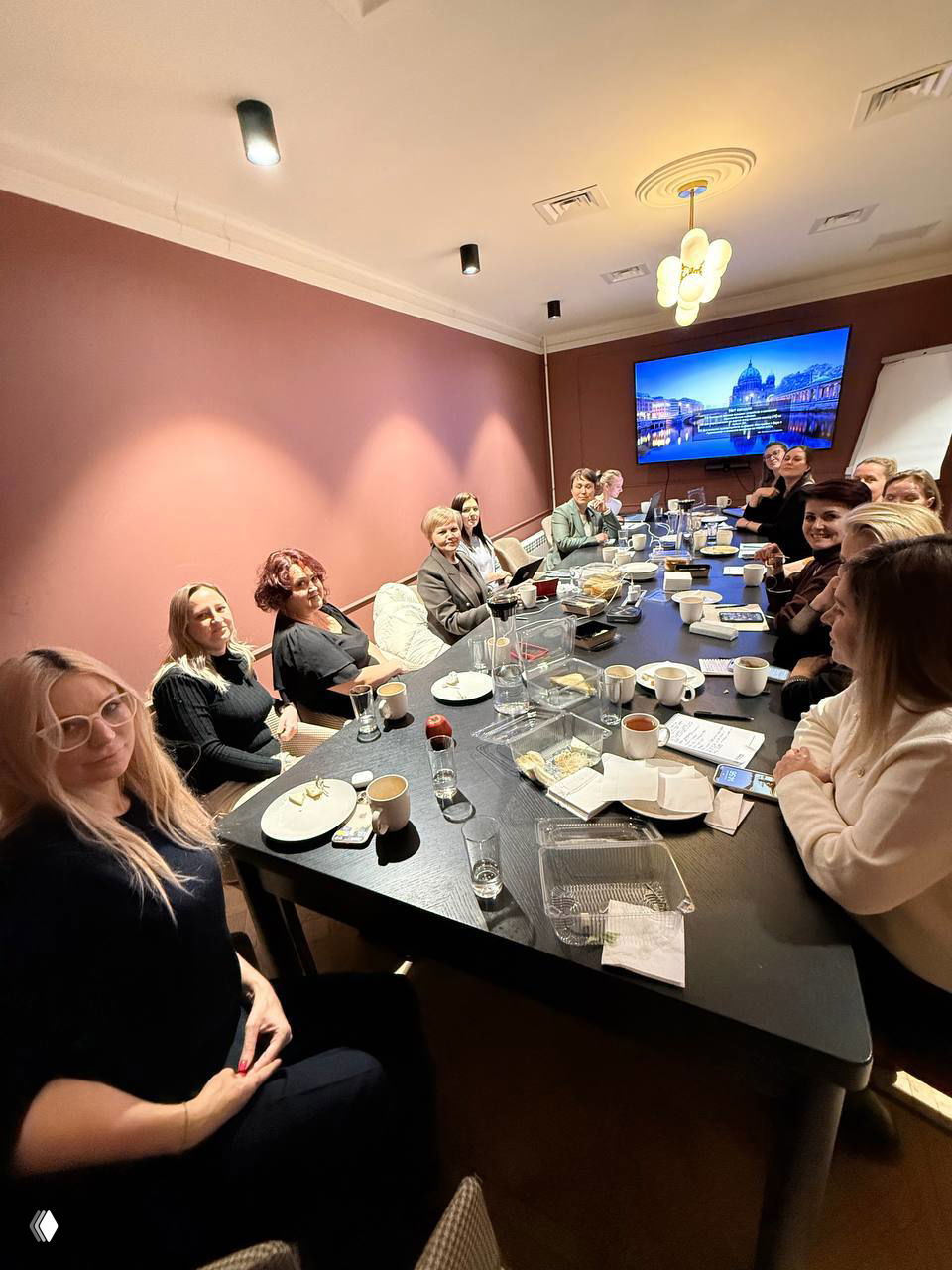 Совещание за длинным столом: несколько женщин с ноутбуками, чашками и блокнотами — рабочая встреча в уютной переговорной.