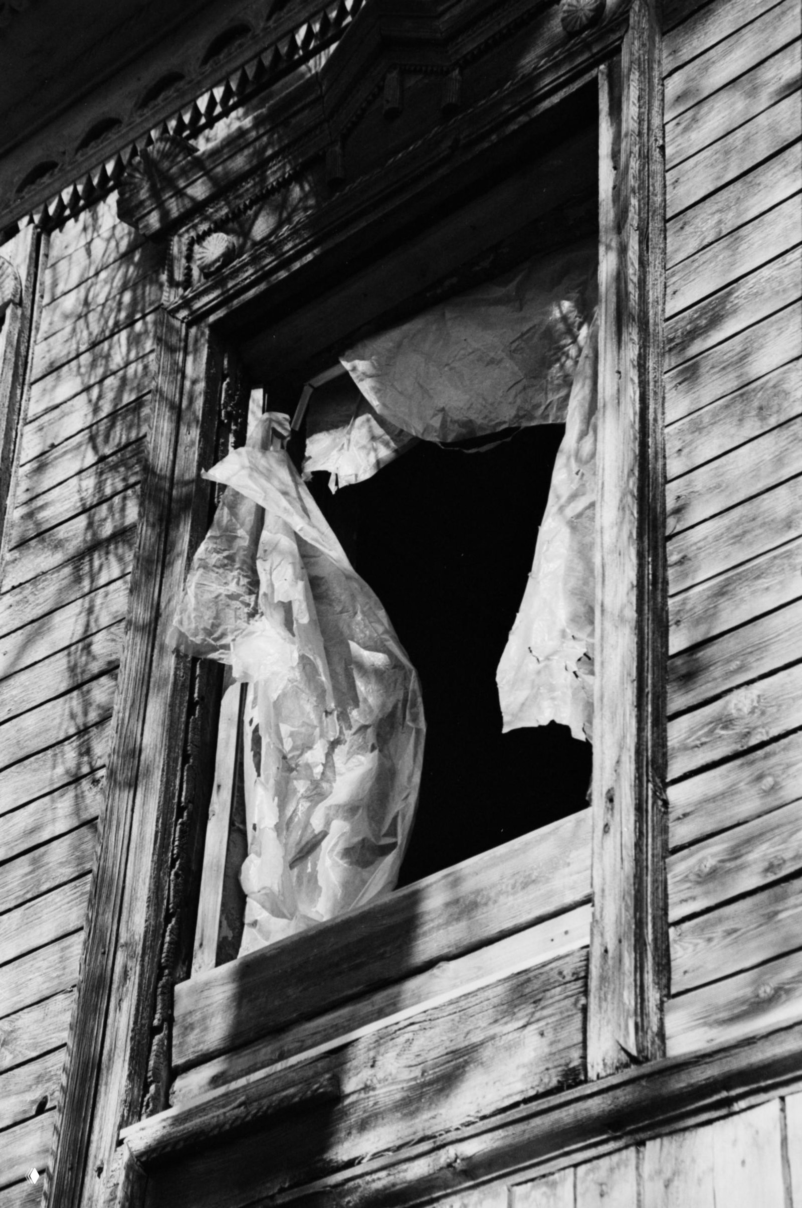 Чёрно‑белая фотография крупным планом: порванная плёнка или пакет в оконном проёме старого деревянного дома, выраженная текстура древесины и резкие контрасты.