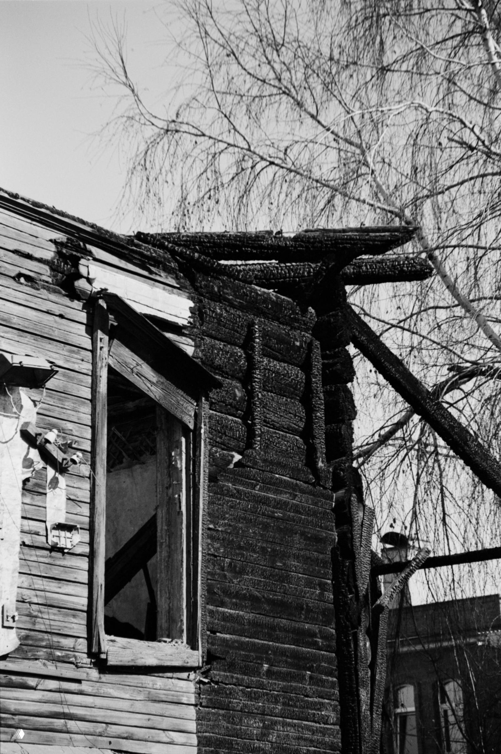 Чёрно‑белый кадр фасада и угла горевшего деревянного дома: обугленные балки, открытое пустое окно и силуэт берёзы на фоне неба, контрастная текстура.