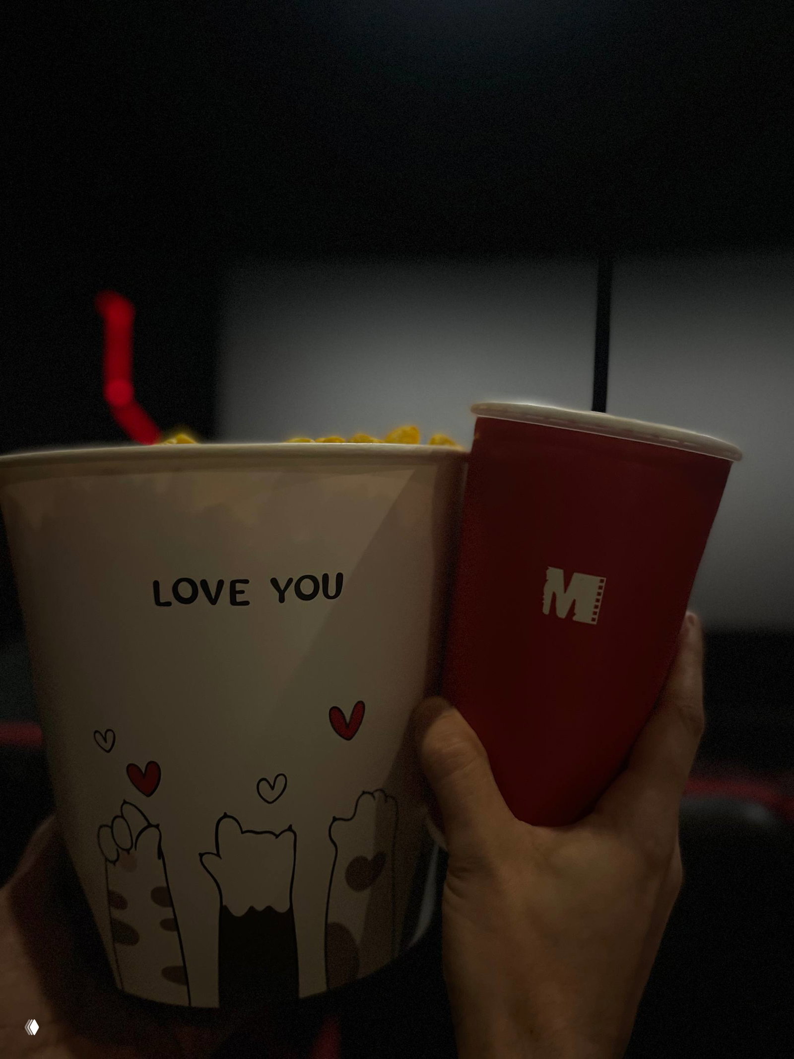 Снимок из зрительного ряда: большая коробка попкорна с надписью «LOVE YOU» и бумажный стакан с напитком в руках зрителя.