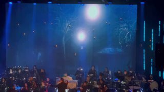 Кадр сцены крупным планом: музыканты оркестра и экран с киноматериалом, освещение, партитуры на пультах музыкантов.