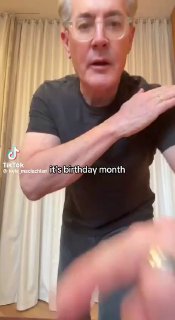 Мужчина в тёмной футболке танцует перед шторами — вертикальный кадр из короткого ролика в TikTok, в очках и с улыбкой.