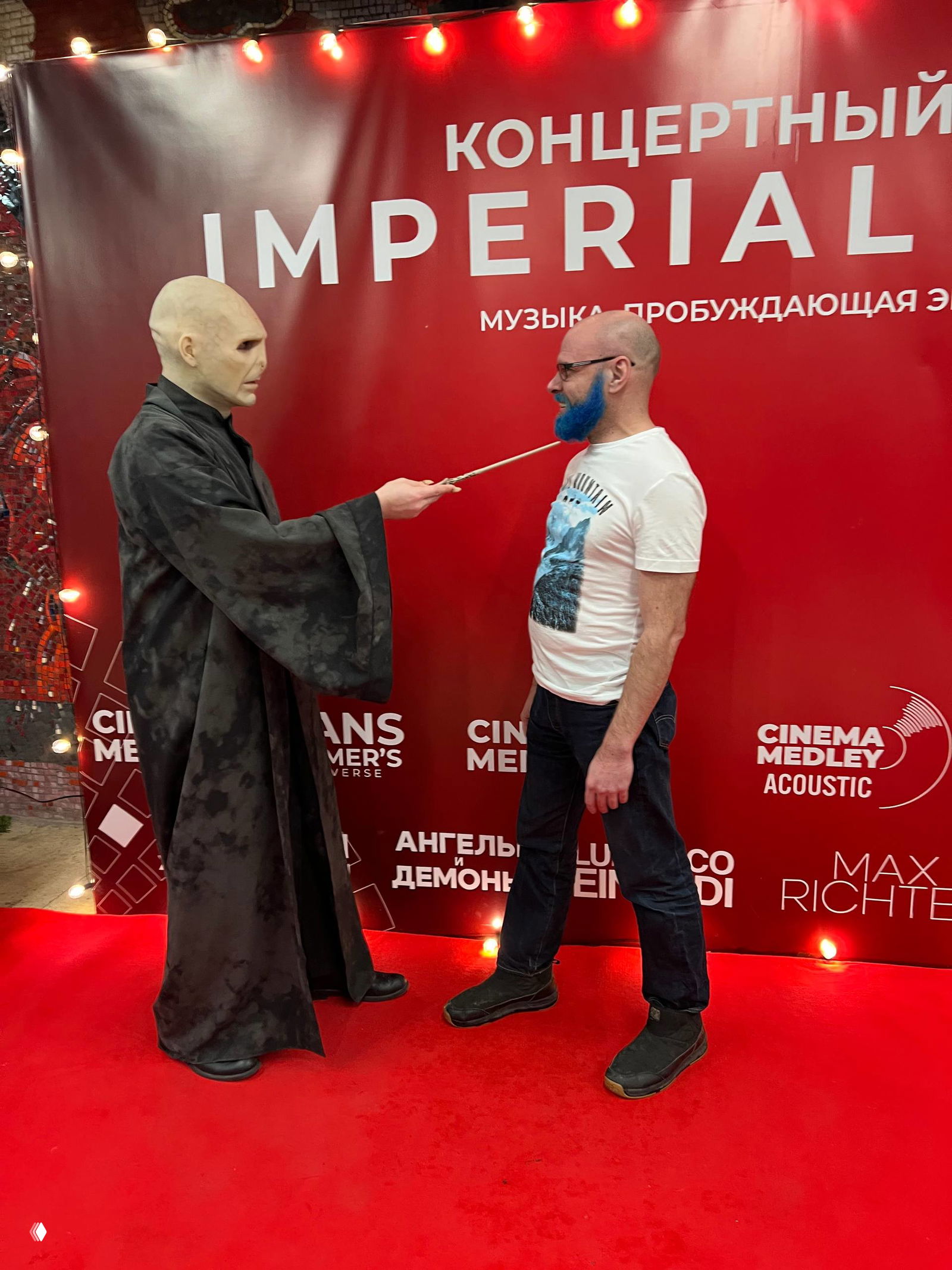 Косплеер Воландеморт в костюме касается волшебной палочкой посетителя с синим бородой на красной дорожке у брендированной фотозоны.