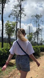 Вид сзади: автор идёт по тропе в белой бланковой футболке и джинсовой юбке, показана длина изделия и прямой крой на фоне деревьев и неба