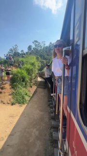 Автор выглядывает из окна поезда в Шри-Ланке, поезд идёт по живописному маршруту через холмы, люди и природа вокруг видны сбоку.