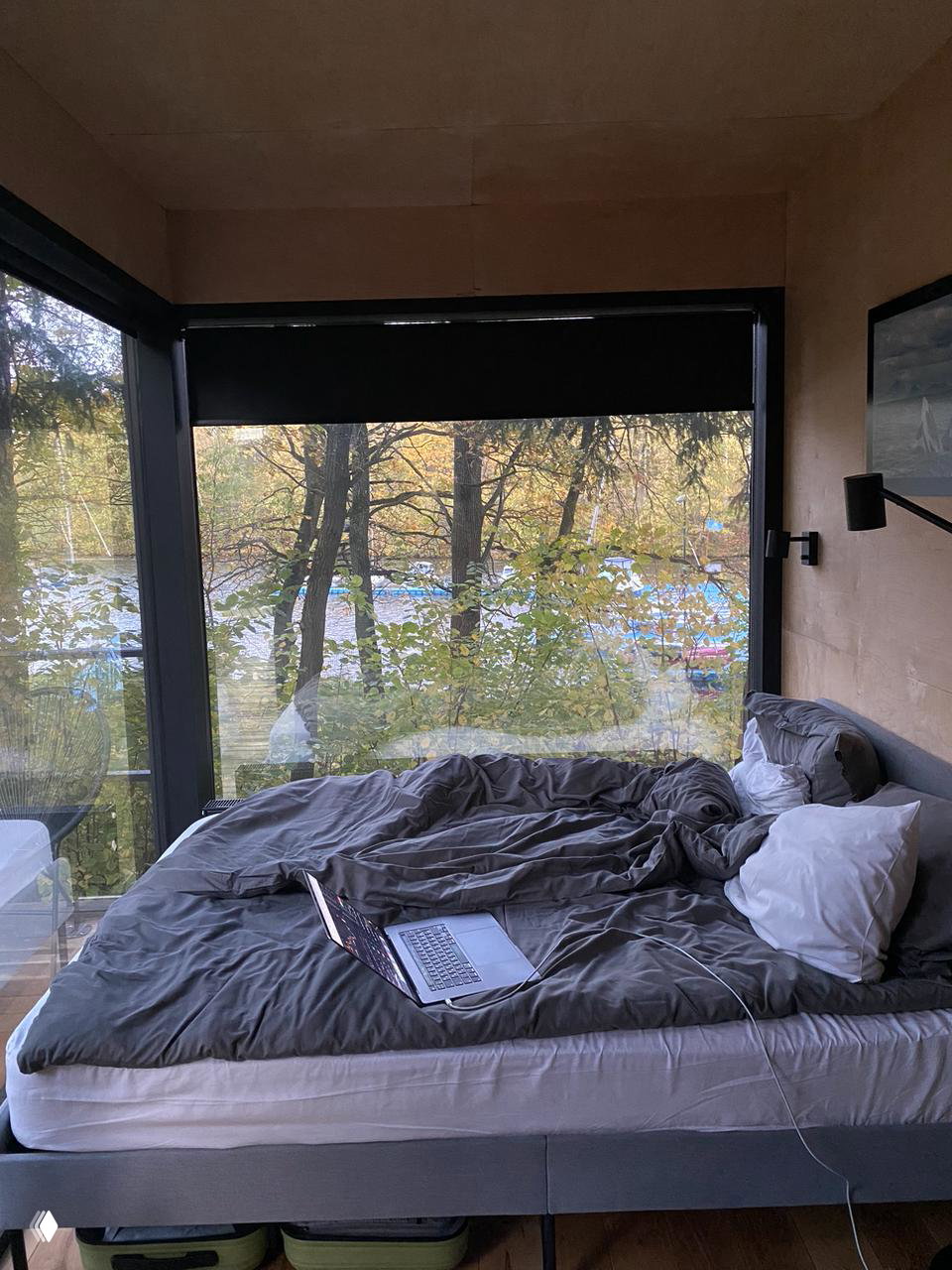 Интерьер глэмпинга с большой панорамной стеной: уютная кровать, ноутбук на простыне и вид на лес и реку за окном, спокойная атмосфера.