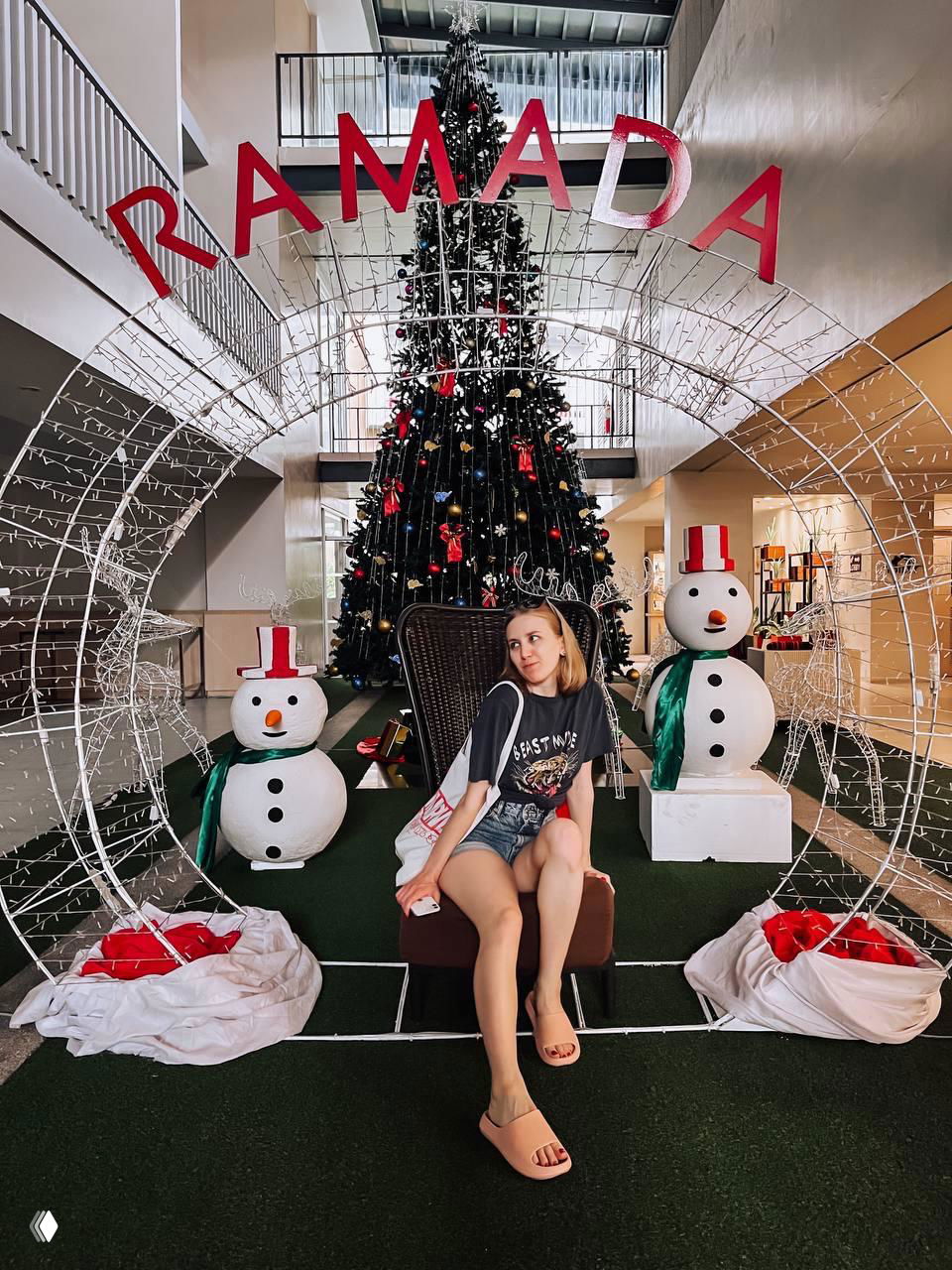 Лобби отеля с большой новогодней ёлкой и праздничными декорациями: девушка сидит на кресле у ёлки, праздничная сценография в Ramada.