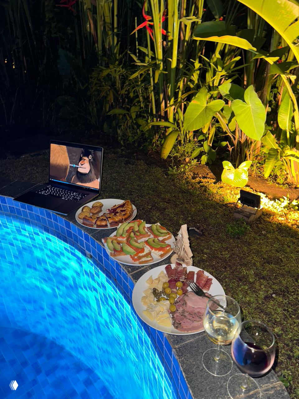 Ночной стол у бассейна: тарелки с закусками, бокалы вина и ноутбук на краю освещённого бассейна, тропические растения вокруг — вечер у бассейна.