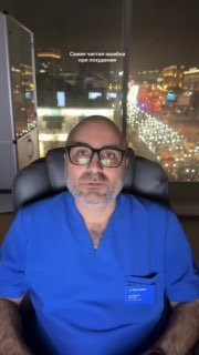 Врач в синем медицинском халате сидит за рабочим столом вечером у окна; через стекло видна ночная городская улица с огнями.