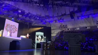 Сцена и ведущий на фоне большой проекции дерева, часть зала заполнена зрителями, общая фиолетово-голубая подсветка и элементы декора видны ясно.