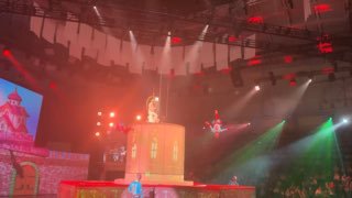 Фрагмент представления: артисты и акробаты в прыжке над сценой, яркие разноцветные прожекторы и красочные сценические декорации в кадре.