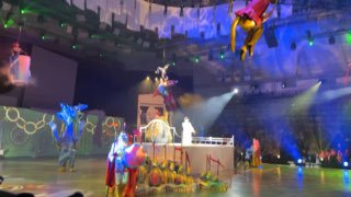 Воздушные артисты парят над сценой, видны крупные сценические конструкции, сложное световое оформление и зрители по краям арены.