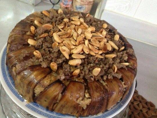 Перевёрнутая сирийская маклюба на блюде: слои риса, жареных баклажанов и фарша, сверху золотистые орешки; домашняя подача.