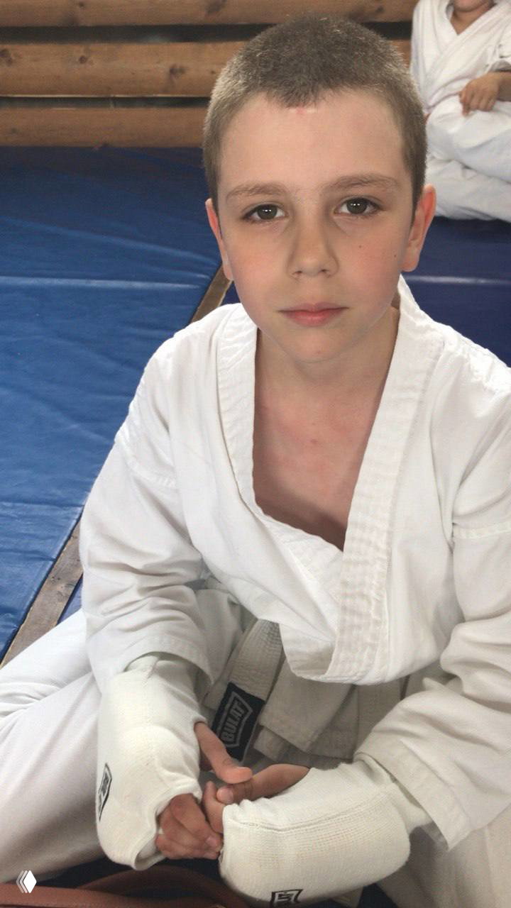 Мальчик в кимоно сидит на татами после тренировки; на руках защитные бинты, серьёзный взгляд в камеру, спортивная атмосфера.