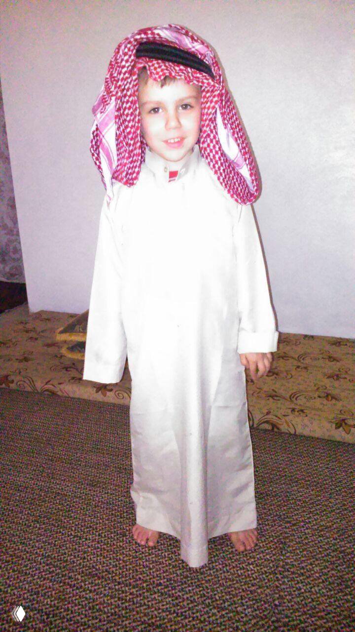 Ребёнок в национальном костюме стоит в комнате; улыбающееся лицо и детская поза, интерьер дома на заднем плане.