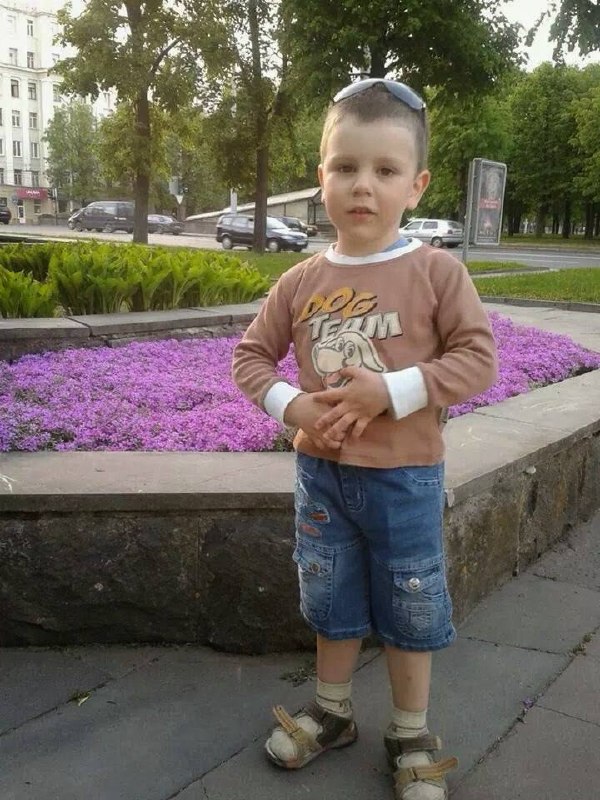 Маленький мальчик стоит у цветочной клумбы в городской среде; летняя одежда и солнечная прогулка по городу, видна улица и автомобили.