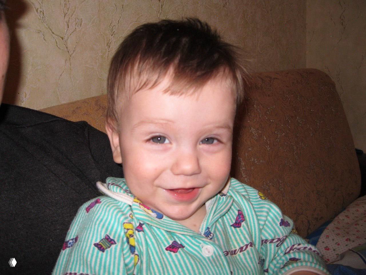 Маленький ребёнок улыбается в домашней обстановке; крупный план лица с искренней детской улыбкой и домашним фоном.