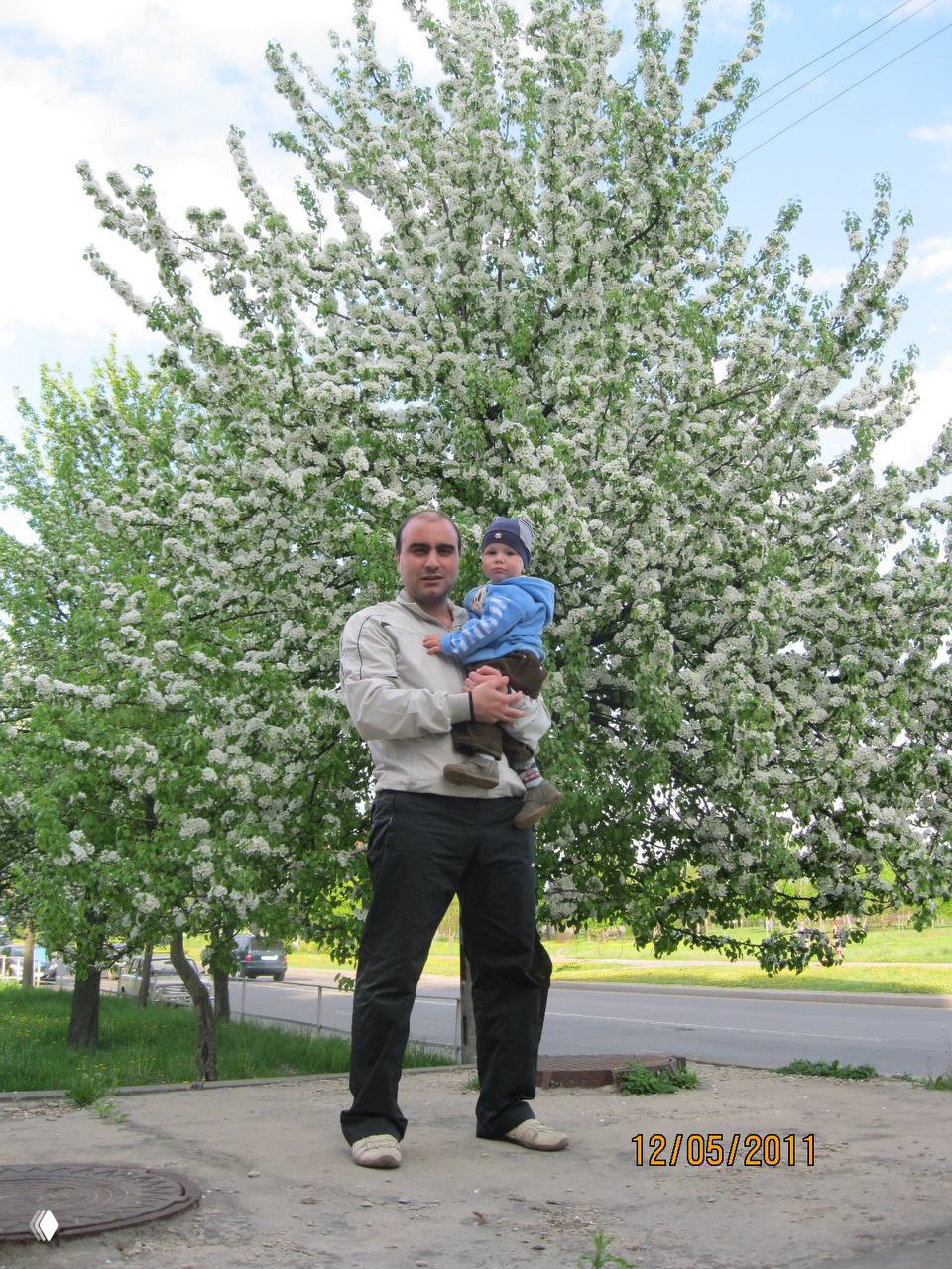 Отец держит младенца на руках на фоне цветущего дерева; весенняя сцена на улице, папа и ребёнок вместе в семейном кадре.