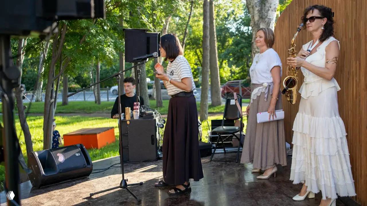 Боковой план сцены с диктором у микрофона, поэтесса и саксофонистка стоят рядом — атмосферный кадр вечернего паркового концерта.