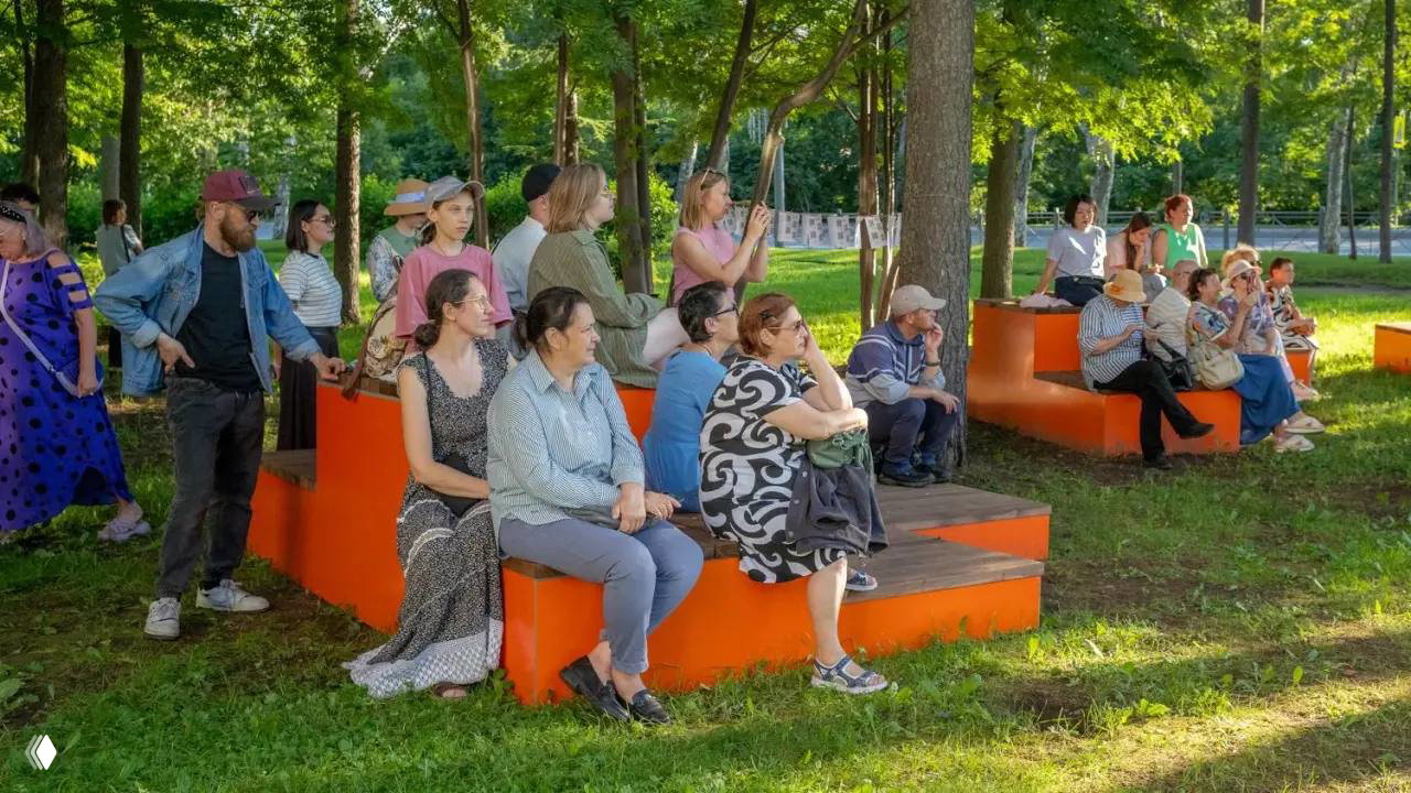 Зрители на оранжевых лавочках в парке внимательно слушают выступление — публика фестиваля «Городская симфония» наслаждается вечером.