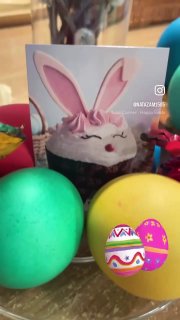 Праздничная композиция: пасхальный кулич с наклейкой в виде зайчика на фоне нескольких цветных крашеных яиц на столе в домашней обстановке.