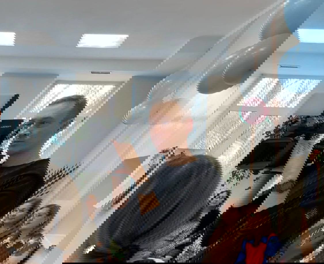 Оператор с профессиональной камерой снимает мероприятие в роддоме, на заднем плане сидящая аудитория и праздничные шары — документирование праздника.