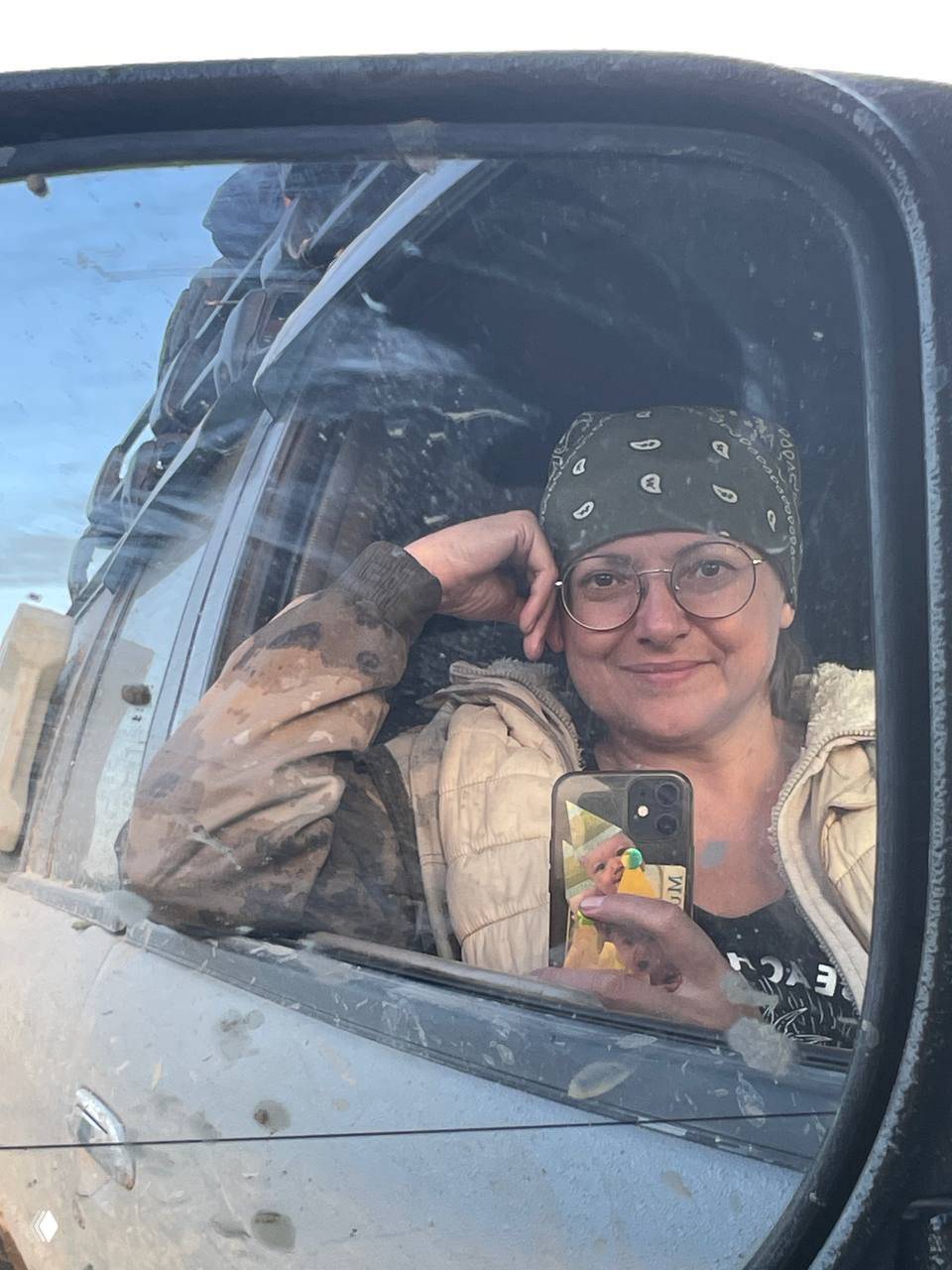 Отражение автора в боковом зеркале внедорожника: женщина в бандане улыбается, на стекле видны следы грязи и салон автомобиля.