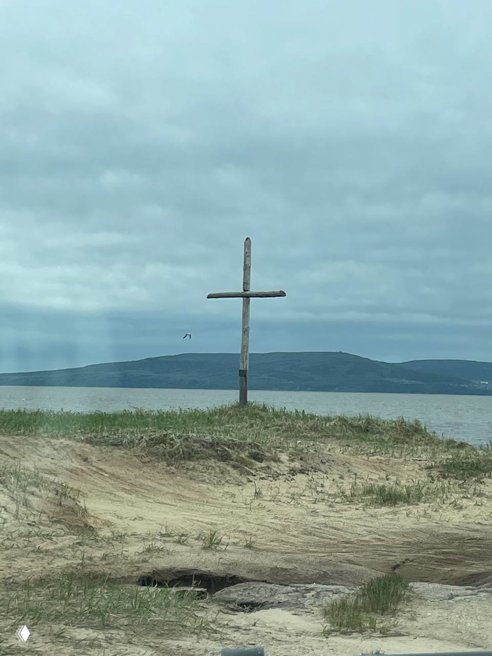 Деревянный крест на песчаном мысе на фоне моря и удалённой горы материка — панорама мыса Погиби с видом на пролив.