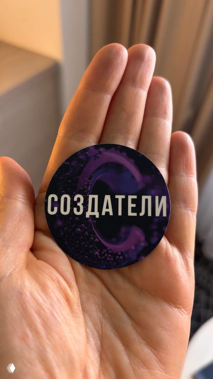 Крупный план: круглый стикер с надписью «СОЗДАТЕЛИ» в руке; фиолетовый дизайн эмблемы и текстура поверхности видны в кадре.