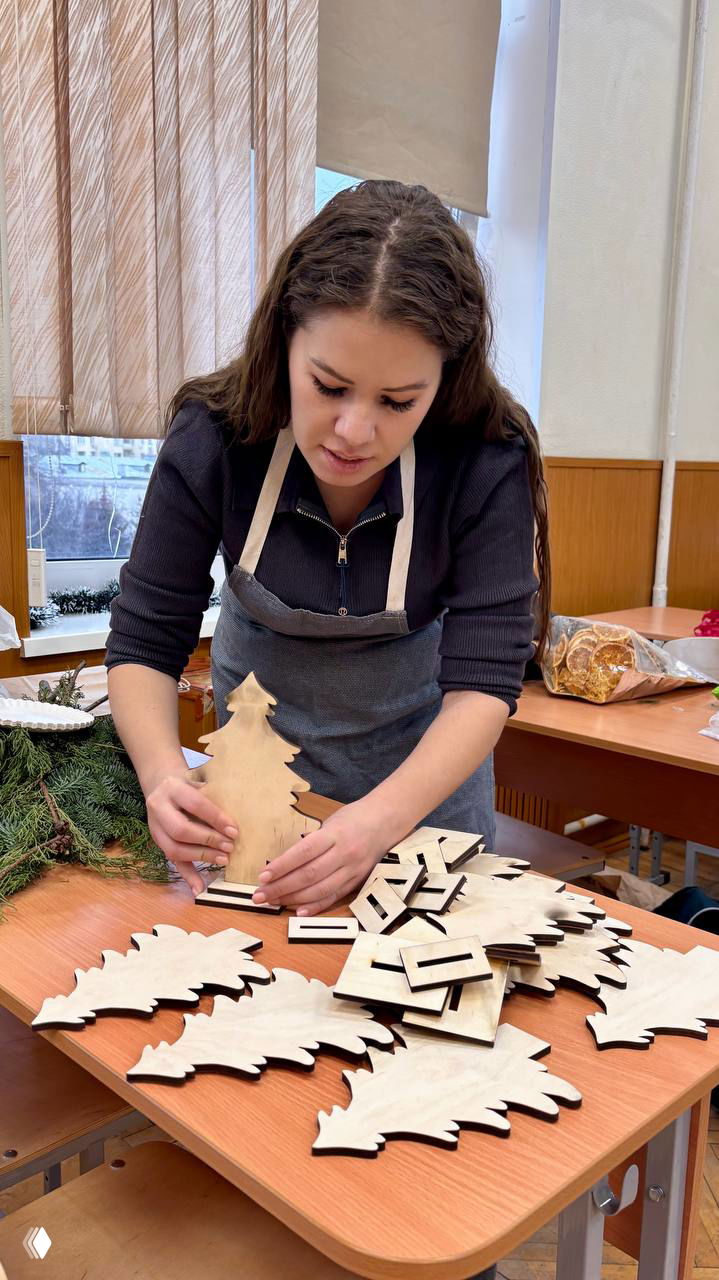 Ученица в фартуке собирает деревянные ёлочки на столе; стопка заготовок и натуральный декор рядом.