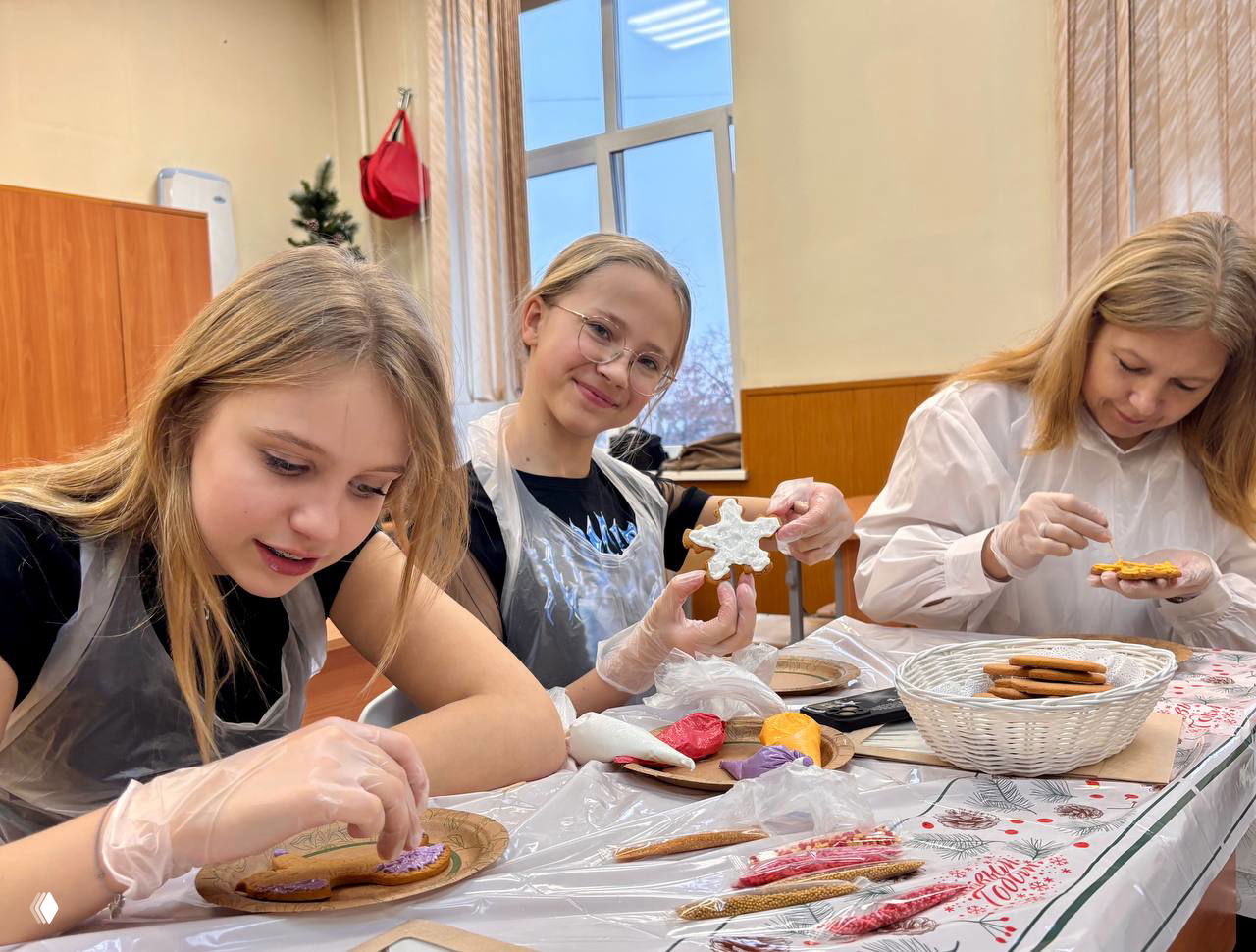 Три девочки за столом расписывают пряники в перчатках; на столе корзина с печеньем и разноцветная глазурь.