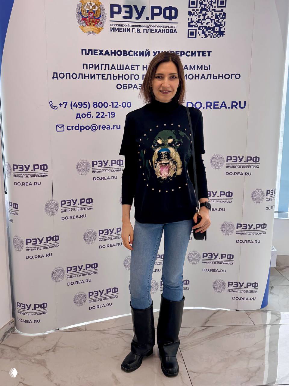 Автор у бренд-стенда Плехановского университета на Дне открытых дверей в легкой одежде, фотография в холле с информацией о программах.