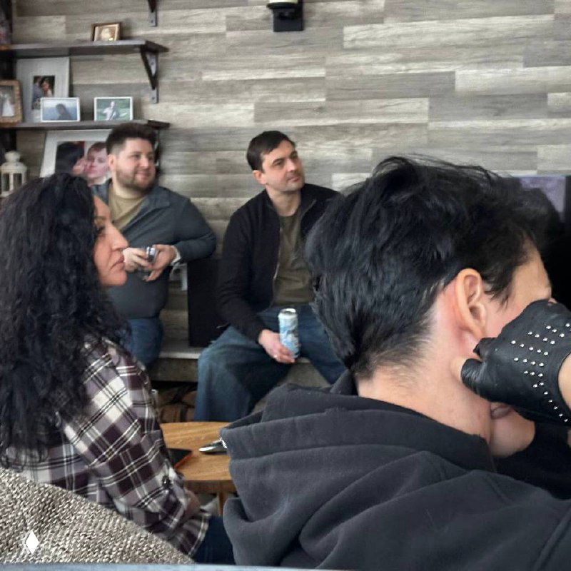 Ещё одна компания гостей в уютной комнате: слушают выступающего, держат напитки, непринуждённая и тёплая обстановка встречи.