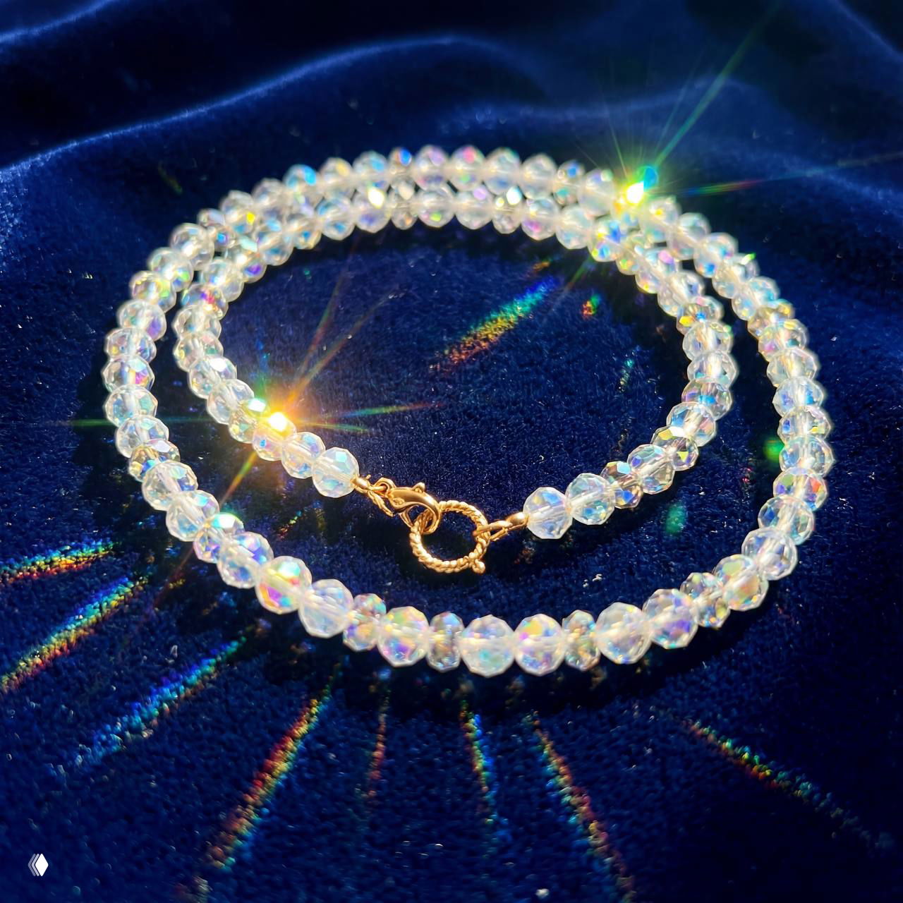 Колье из прозрачных радужно сверкающих бусин с золотой застёжкой на синем бархатном фоне; блики света создают праздничную атмосферу.