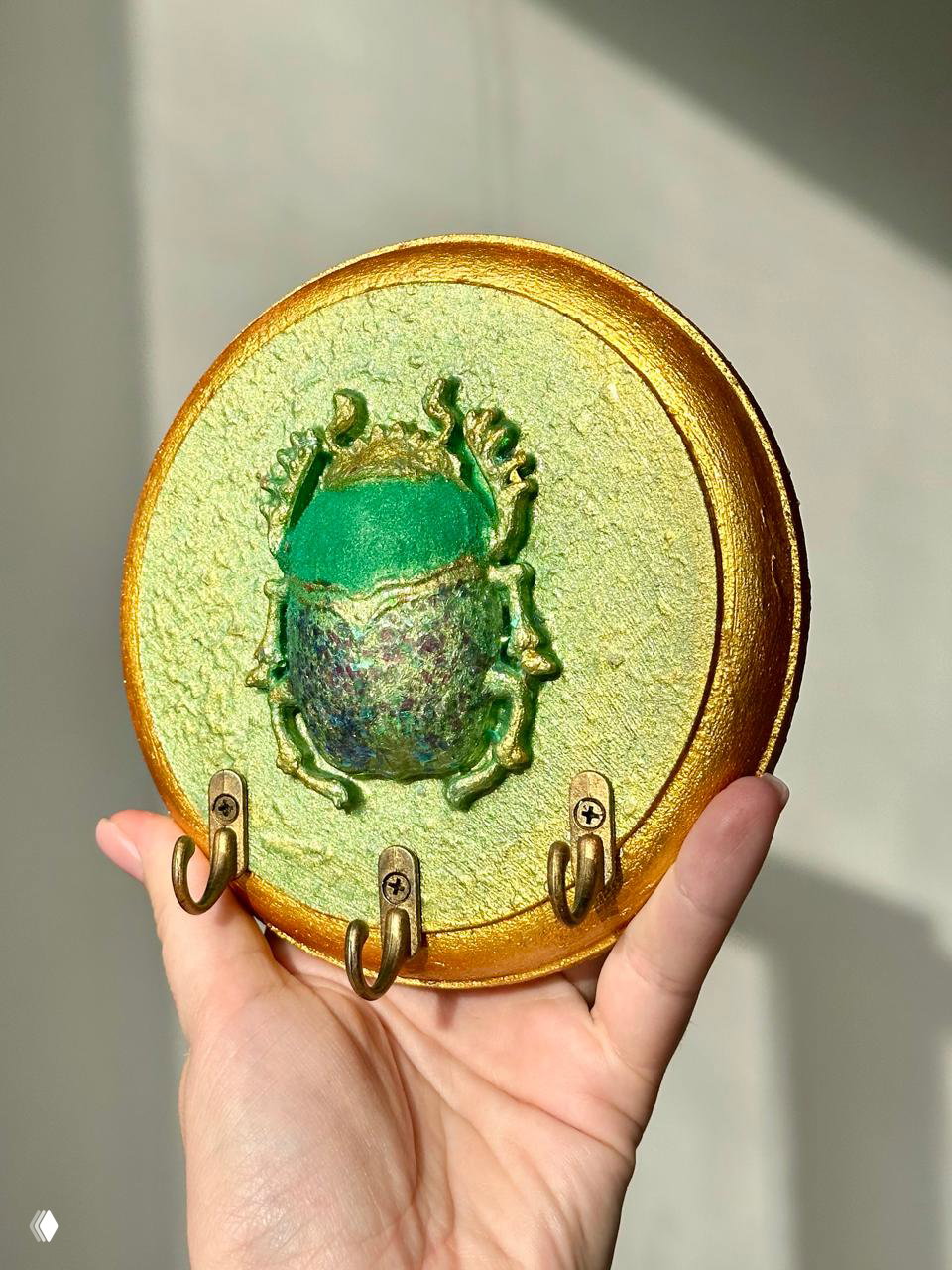Портретный план круглой ключницы с выпуклым зелёным жуком и тремя крючками, видна фактура краски и золотистая рамка на светлом фоне.