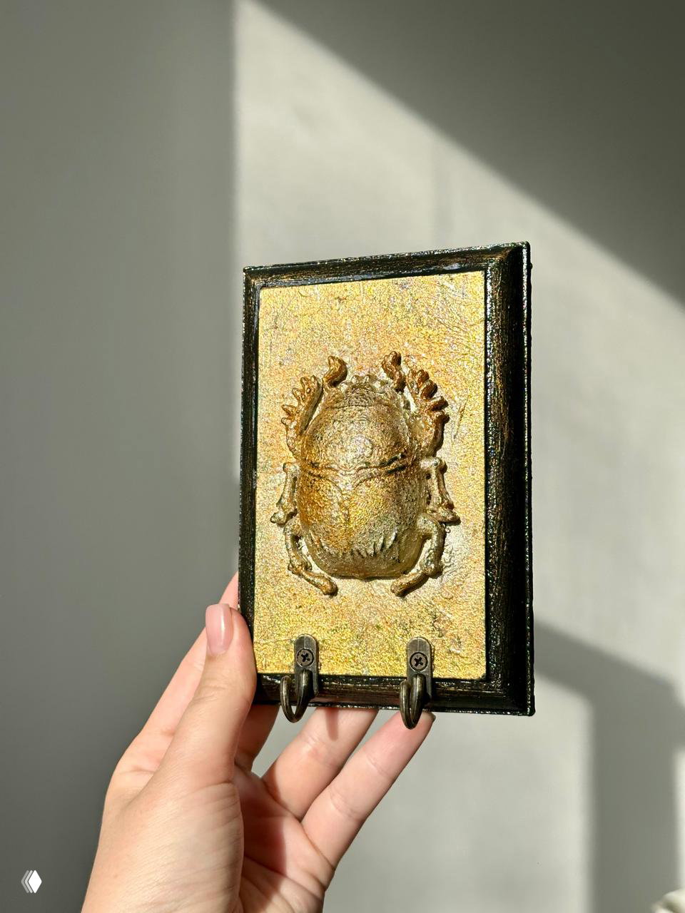 Прямой кадр вертикальной прямоугольной ключницы с объёмным золотистым жуком на текстурированном фоне в тёплом освещении.