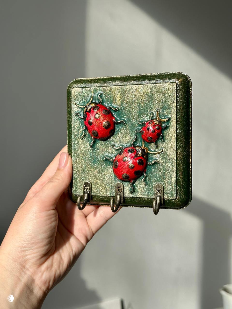 Квадратная ключница с тремя объёмными красными божьими коровками на зелёно‑золотистом фоне, рука держит изделие на сером фоне.