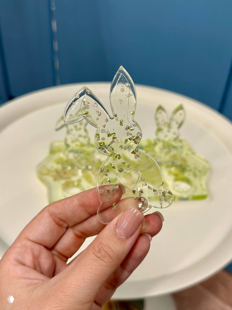 Рука держит прозрачную фигурку‑зайчика из эпоксидной смолы с блёстками на фоне подставки; демонстрация масштаба и качества обработки.