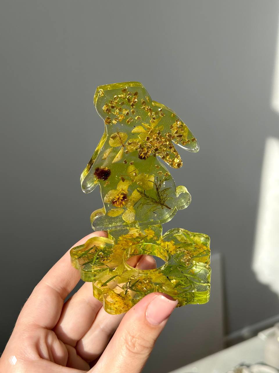 Фото желто-зелёной подставки для яйца из эпоксидной смолы с вкраплениями растений и золотистого декора, показано на свету в руке.