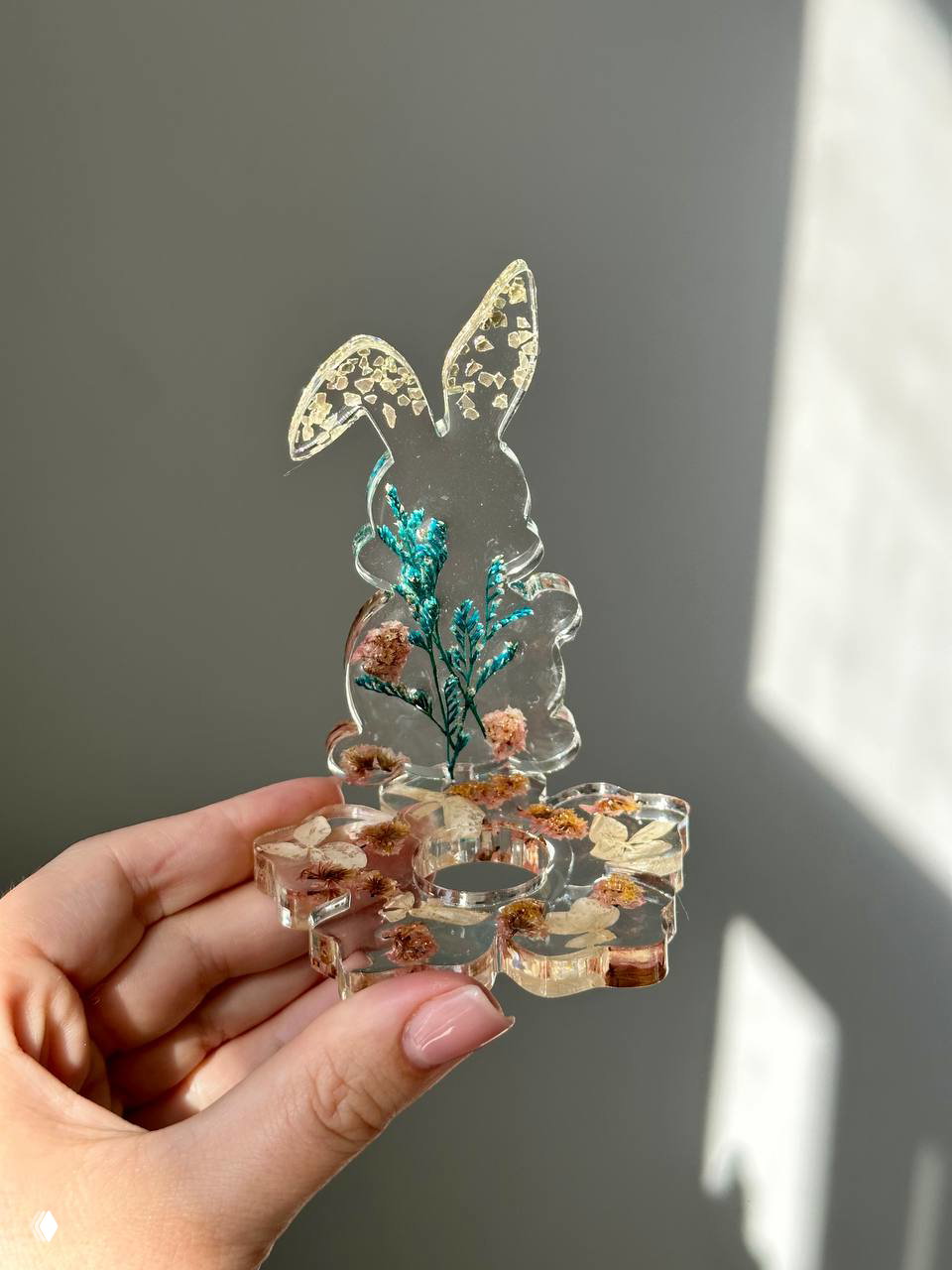 Прозрачная подставка с фигуркой зайца из эпоксидной смолы, внутри видны сухие цветы и бирюзовые акценты, фон — светлая стена.