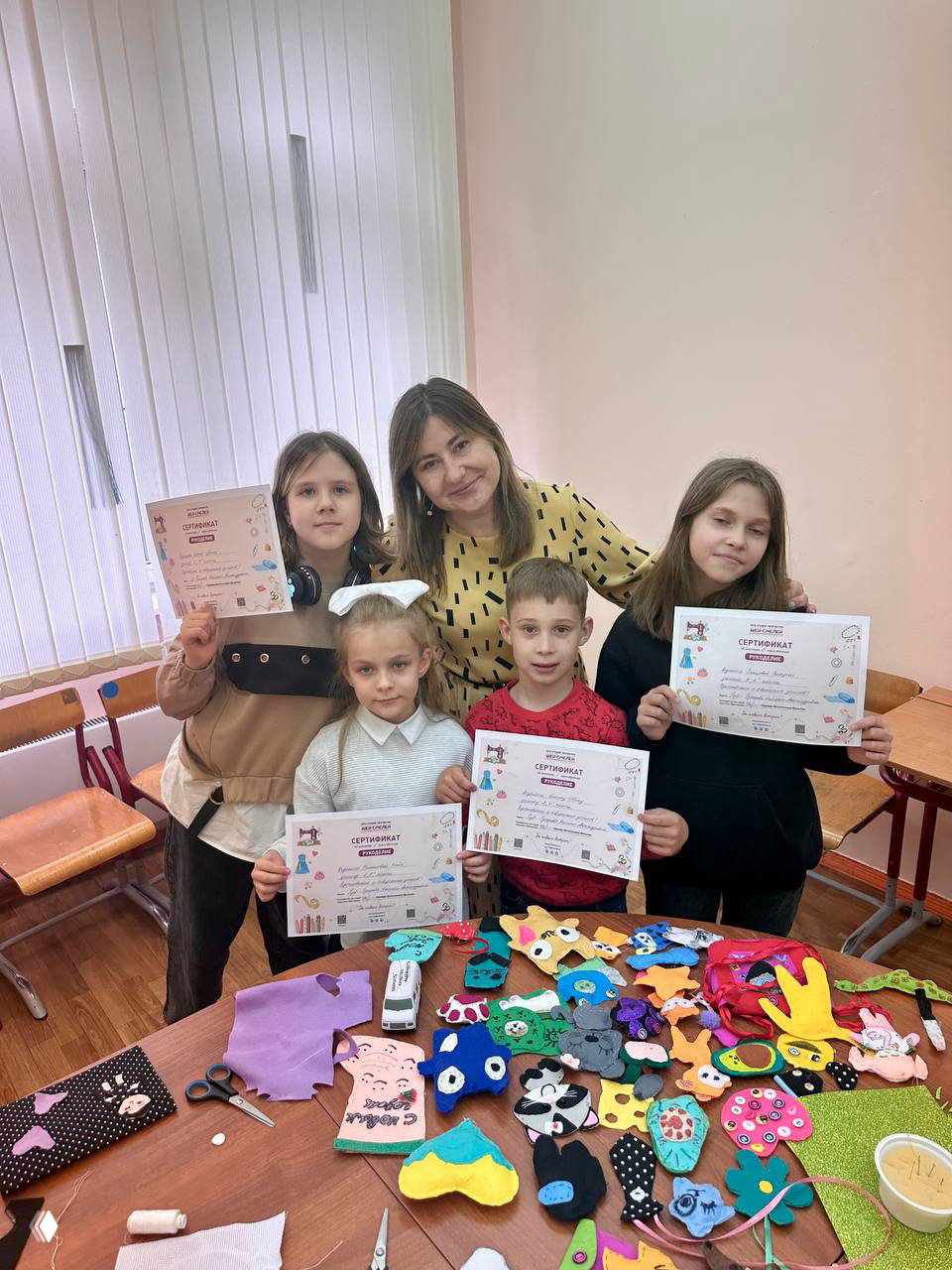 Преподаватель с группой детей показывает сертификаты; на столе яркие фетровые игрушки, аппликации и готовые изделия после мастерской.