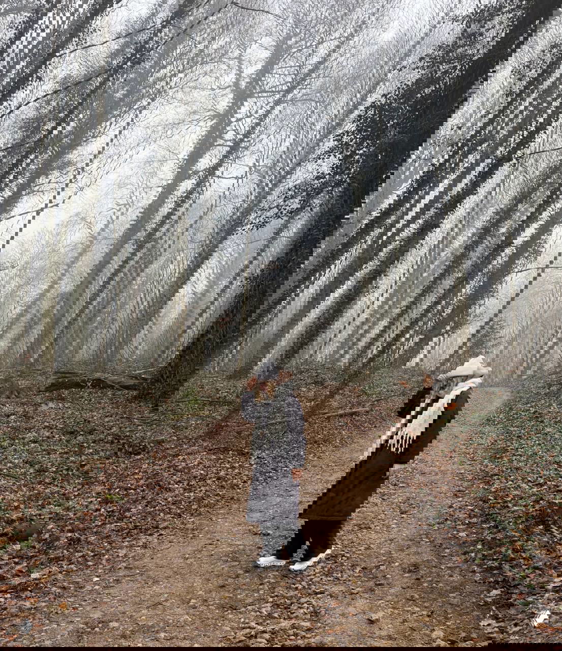 Авторка прогулки в тёплом пальто и шапке идёт по лесной тропе среди высоких деревьев; сцена подчёркивает спокойную атмосферу окрестностей города.