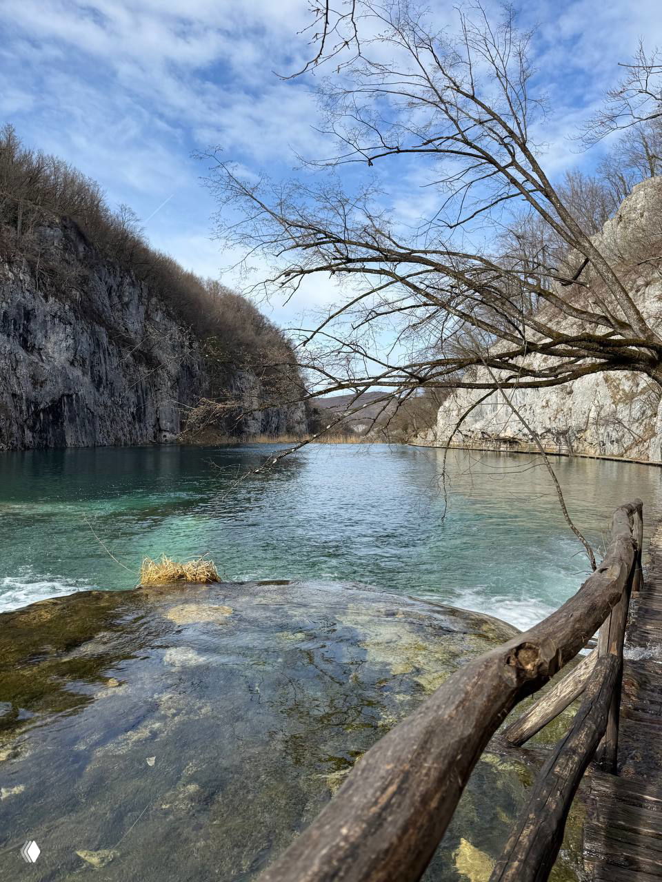 Спокойная гладь озера между крутыми скалами и голыми деревьями в начале сезона; деревянное ограждение и прогулочная тропа на переднем плане.