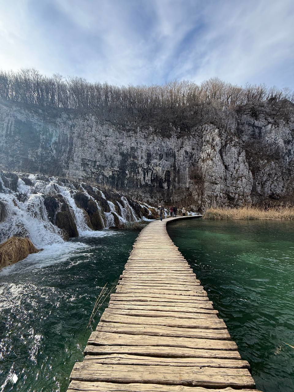 Деревянный настил, ведущий через прозрачную воду к скалам и каскадам; узкая тропинка вдоль водопадов с туристами на фоне.
