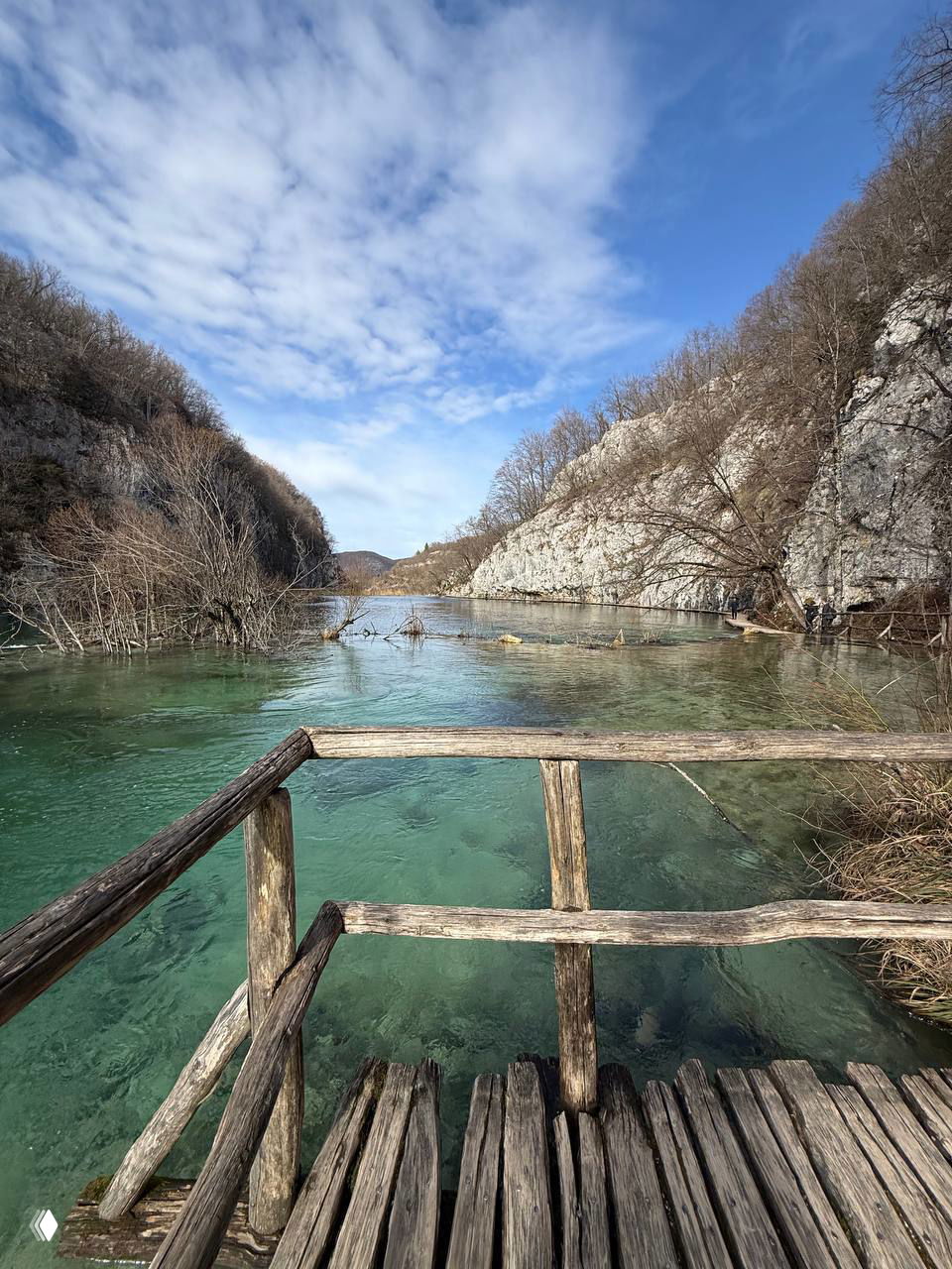 Прозрачная бирюзовая вода и деревянная смотровая площадка с простым перилом; вид на озеро и окружение из скал и редкого леса.