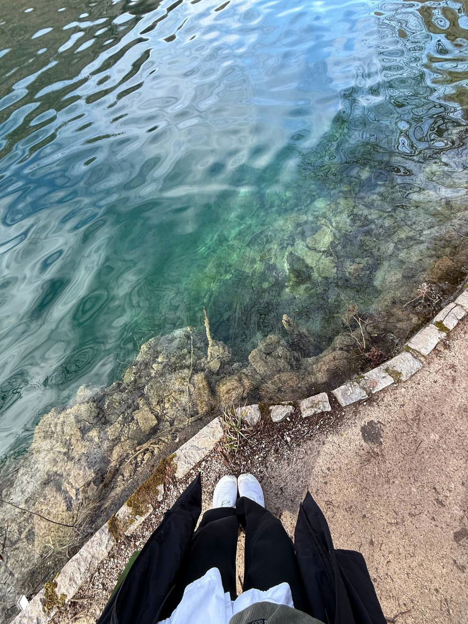 Крупный план прозрачной воды: видны камни и растительность под поверхностью, на переднем плане обувь туриста у кромки тропы.