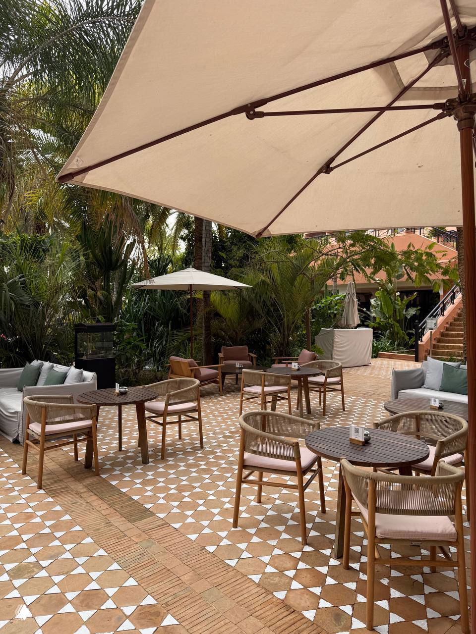 Открытая терраса кафе у отеля в Марракеше: столики и плетёные кресла под большими зонтиками на характерной теракотовой плитке среди пальм.