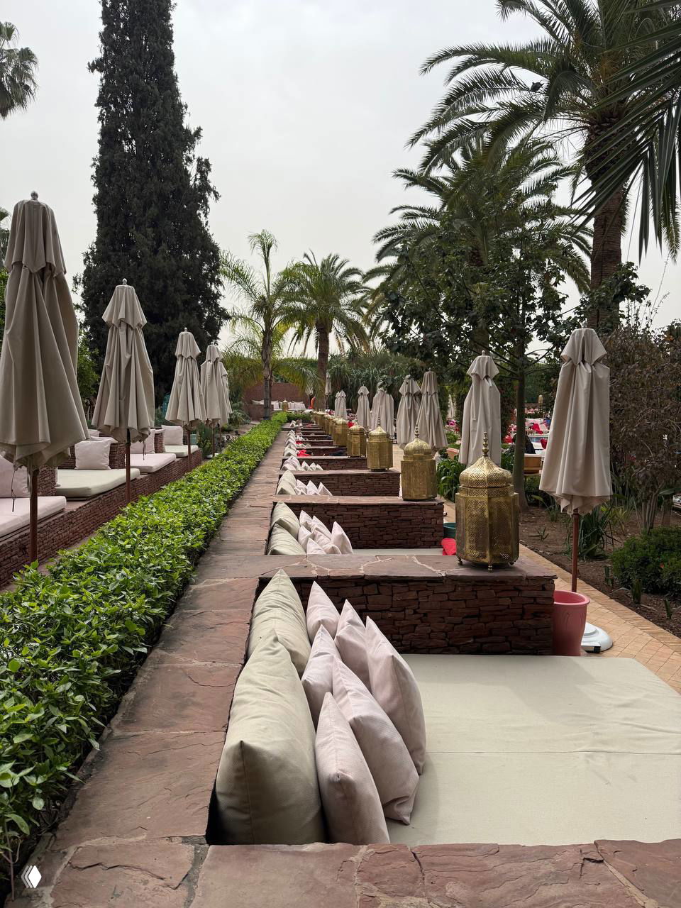 Ряд вкопанных ложей с подушками вдоль зоны отдыха у бассейна в марокканском стиле: фонари, зонтики и уютная расстановка для релакса.