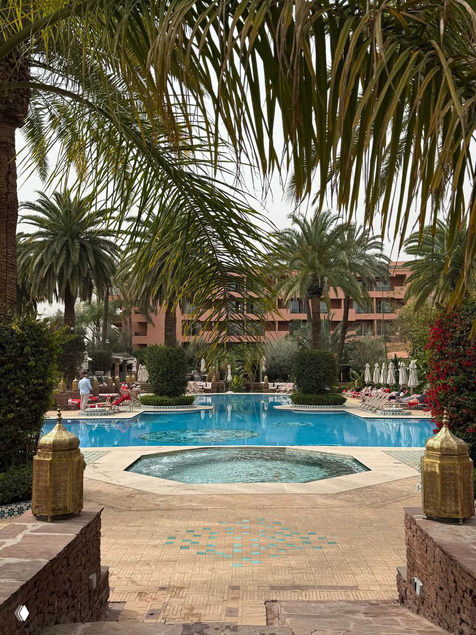 Вид на большой открытый бассейн отеля с пальмами и декоративным джакузи на переднем плане, мозаика и архитектура в марокканской стилистике.